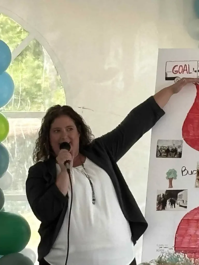 Harmony Centre Executive Director Jennifer speaking at a fundraising celebration, pointing to a campaign goal poster surrounded by balloons.