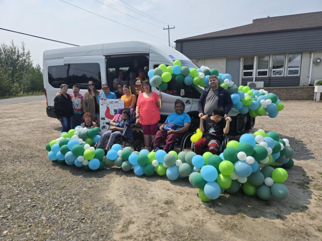 Harmony Centre participants and staff celebrating outdoors beside a new accessible van, surrounded by balloon arches and a fundraising goal sign.